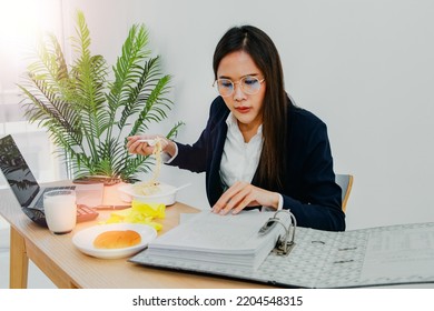 Asian Businesswoman Dressed Suit Early In The Morning, Rushes To Clear Business Papers In Her Home Office, Sits Down And Eats Ready To Eat Breakfast Of Milk And Bread Before Leaving For Work.
