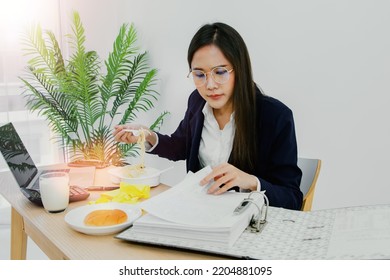 Asian Businesswoman Dressed Suit Early In The Morning, Rushes To Clear Business Papers In Her Home Office, Sits Down And Eats Ready To Eat Breakfast Of Milk And Bread Before Leaving For Work.
