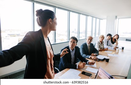 Asian Businesswoman Explaining New Business Strategy To Coworkers Sitting Around Table In Conference Room. Businesspeople During Presentation In Board Room.