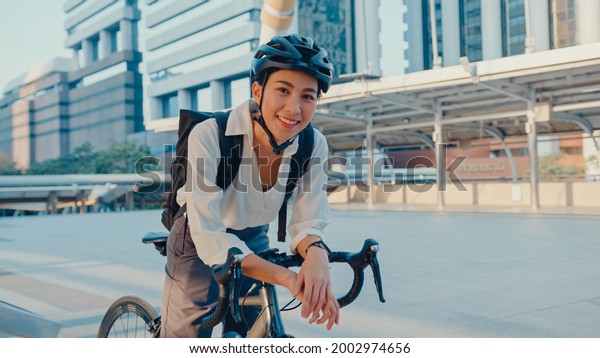 Asian businesswoman go to work at office stand\
and smiling wear backpack look at camera with bicycle on street\
around building on a city. Bike commuting, Commute on bike,\
Business commuter\
concept.