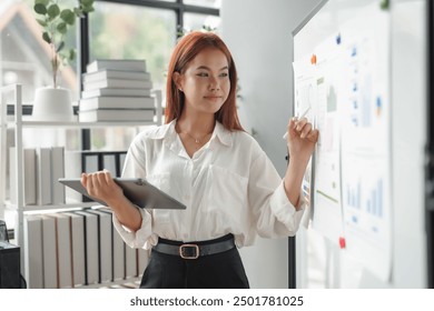 Asian businesswoman is holding a digital tablet and pointing at a whiteboard with charts and diagrams - Powered by Shutterstock