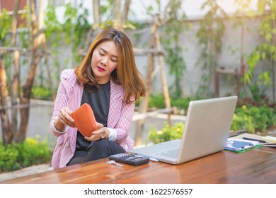 asian businesswoman holding a wallet taking money or credit card out of it, with computer laptop, calculator, key, clipboard pen, and notebook laid on top of the table while sitting with legs crossed 