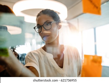 Asian Businesswoman Looking Over A Post It Note Wall And Brainstorming. Mature Woman Standing At The Office Behind Glass Wall With Sticky Notes.