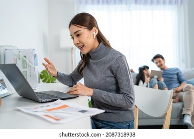 Asian Businesswoman Mother Sit On Table In Living Room, Work From Home. Attractive Mom Use Laptop Computer For Online Meeting In Room With Young Kid Daughter Play With Father On Sofa Together In House