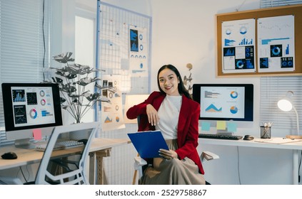Asian businesswoman in a red blazer sits in her office chair in her office, holding a blue clipboard. Charts and graphs are visible on her computer monitors and on the walls