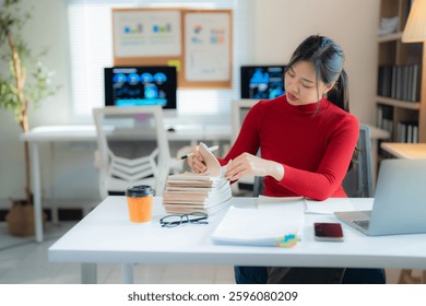 Asian businesswoman in a red turtleneck reviewing notes in her notebook while working diligently at her desk in a modern office, surrounded by technology and data analysis tools