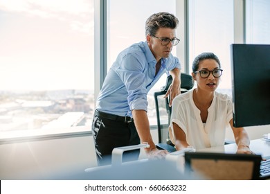 Asian Businesswoman Sitting At Her Desk And Working On Computer With Male Colleague Standing By. Two Corporate Professionals Working Together In Office.