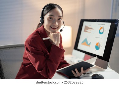 Asian businesswoman is smiling while working late at night in her office. She is wearing a headset and holding a notebook and pen - Powered by Shutterstock