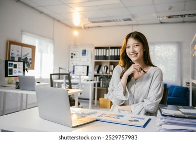 Asian businesswoman smiling and working at her office desk surrounded by paperwork and financial graphs, exuding confidence and professionalism under natural light - Powered by Shutterstock