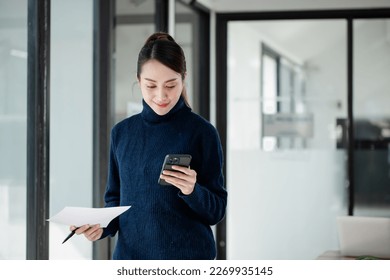 Asian businesswoman standing with paperwork at workplace using smartphone online in a modern office.  - Powered by Shutterstock