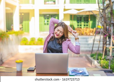 Asian Businesswoman Stretching With Both Of Her Arms Bent Behind Her Back With Back Straight, After Getting Tired From Working On The Laptop Computer, Calculator, Notebook And Clipboard On The Table