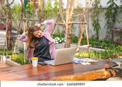 Asian Businesswoman Stretching With Both Of Her Arms Bent Behind Her Back With Back Straight, After Getting Tired From Working On The Laptop Computer, Calculator, Notebook And Clipboard On The Table