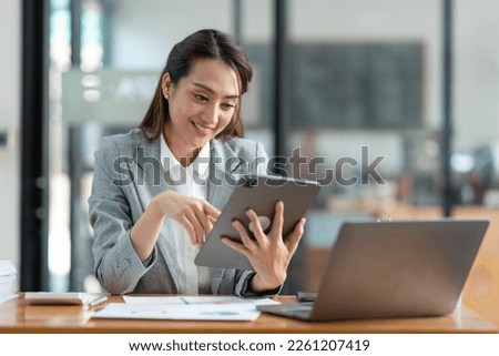 Asian businesswoman in a suit sitting holding iPad or tablet in contact See details and discuss business information happily in the office.
