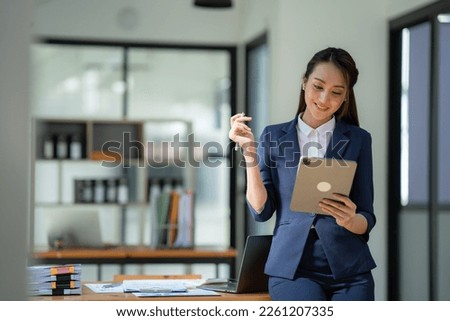 Asian businesswoman in the suit standing confidently holding iPad or tablet in hand and smiling in the office.