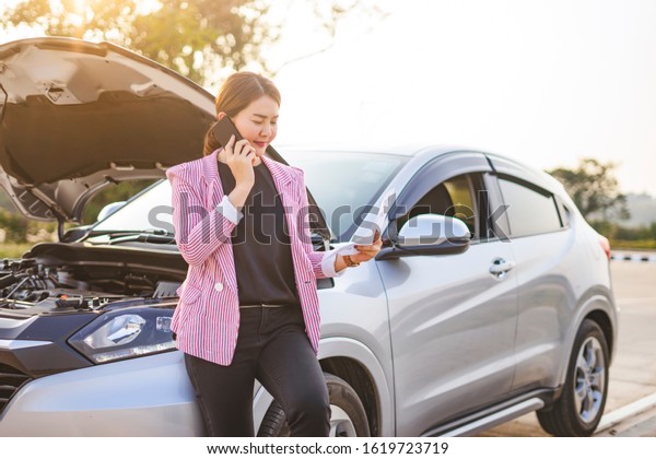 asian businesswoman talking on the phone holding\
a piece of paper reading the information, leaning against a broken\
down car with the hood opened up, with car parking on the side of\
the road highway