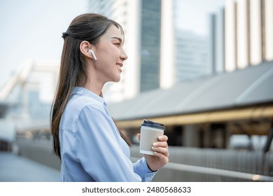 Asian businesswoman talking through wireless headphones while walk in city. Attractive female employee worker feeling happy and relax, holding a cup of coffee takeaway going to the skyscrapers office - Powered by Shutterstock