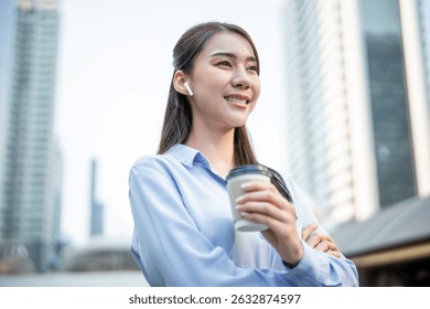 Asian businesswoman talking through wireless headphones while walk in city. Attractive female employee worker feeling happy and relax, holding a cup of coffee takeaway going to the skyscrapers office
