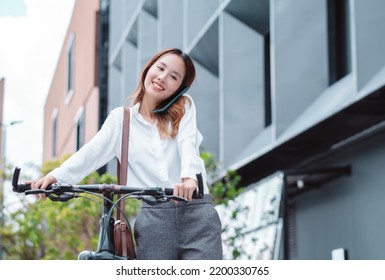 Asian Businesswoman Use Smart Mobile Phone During Go To Office Work At City Street With Bicycle, Happy Female Commuting Outside In Morning, Eco Friendly People Lifestyle Concept.