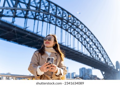 Asian businesswoman using mobile phone during walking city street at Sydney, Australia. Confidence woman enjoy outdoor lifestyle travel Sydney harbour bridge with digital device and online network. - Powered by Shutterstock