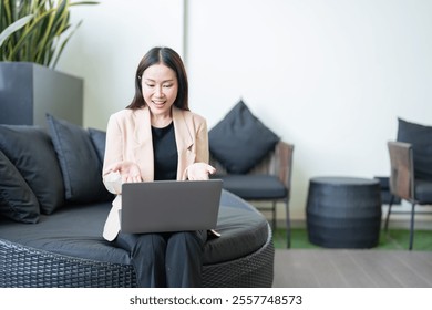 Asian businesswoman wearing beige jacket and black shirt using laptop for video call, gesturing with hands and smiling, sitting on black round sofa in modern office lounge - Powered by Shutterstock