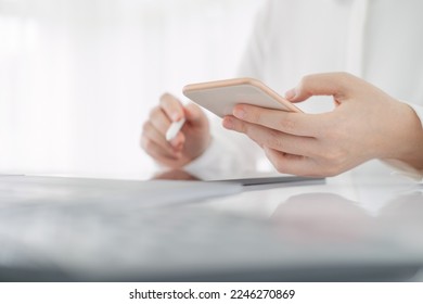 Asian businesswoman in white formal clothes writing note in tablet and checking smart phone. Business woman busy working with documents and tablet  in workstation. close up clean image. - Shutterstock ID 2246270869