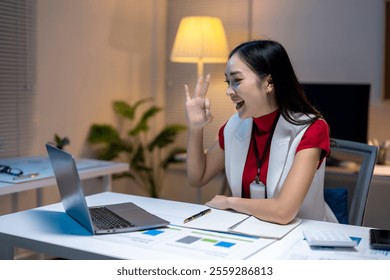 Asian businesswoman working late and making ok sign with hand gesture during video conference call on laptop in office at night - Powered by Shutterstock