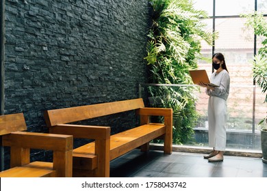 Asian businesswoman working on laptop while standing outside office building. She wears a face mask during covid 19 pandemic. - Full length - Powered by Shutterstock