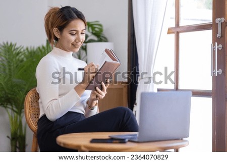Asian businesswoman working on a laptop computer and writing notes at home office, doing calculating expense financial report finance making notes paper graph data document.