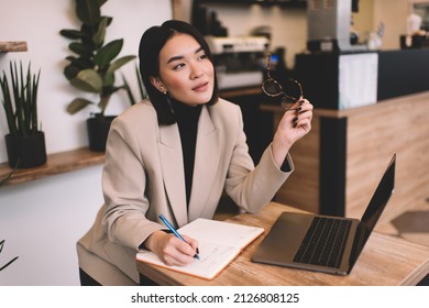 Asian businesswoman writing in notebook and thinking about something in cafe. Young millennial brunette woman sitting at table with opened laptop. Freelance and remote work. Modern female lifestyle - Powered by Shutterstock