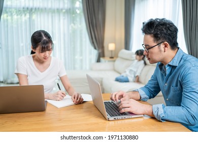 Asian Busy Parents Working From Home Due Covid-19 Lockdown With Children Watching TV Behind. Business Father And Mother Typing Laptop And Write On Copy Book Didn't Pay Attention To Little Daughter.