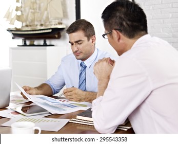 Asian And Caucasian Business Executives Reviewing And Discussing Business Performance In Office Of A Multinational Company.