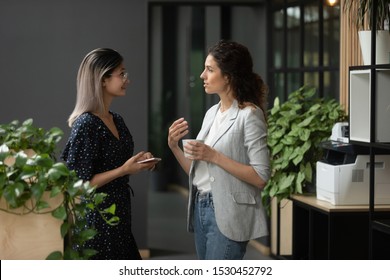 Asian And Caucasian Ethnicity Businesswoman Talking Discuss Common Project Standing In Workplace Hall More Experienced Employee Share Knowledge With Intern During Coffee Break In Modern Office Hallway