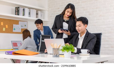 Asian Cheerful Professional Successful Businesswoman Manager Mentor Standing Helping Young Businessman Colleague Sitting Working With Laptop Notebook Computer On Workstation Desk In Company Office.