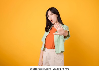 Asian cheerful woman 30s wearing green and orange shirt. With a hand raised in 'no' gesture, expresses conviction isolated on yellow background. - Powered by Shutterstock