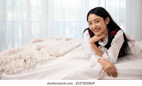 Asian cheerful young woman in casual attire relaxes on cozy bed, smiling warmly in airy bedroom with copy space - Powered by Shutterstock