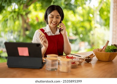 Asian chef woman wearing apron is cooking while having phone call using tablet for online order in kitchen - Powered by Shutterstock