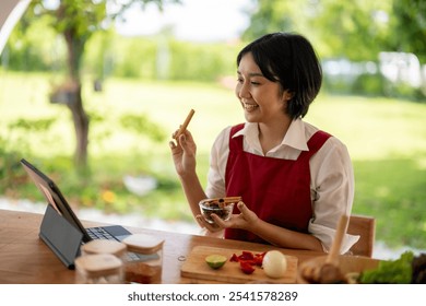 Asian chef woman wearing apron, using laptop, preparing ingredient, cooking while having video call on digital tablet in kitchen garden at home - Powered by Shutterstock