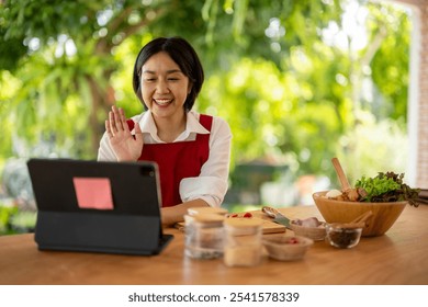 Asian chef woman wearing apron making video call on tablet and waving to someone while preparing ingredients for cooking food in the kitchen at home - Powered by Shutterstock