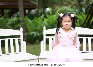 Asian Child Cute Or Kid Girl Smile Sit On White Wood Chair For Relax In Green Garden At Home On Wedding Ceremony For Bridesmaid And Wear Pink Princess Or Fairy Costume Or Dress On Love Valentine