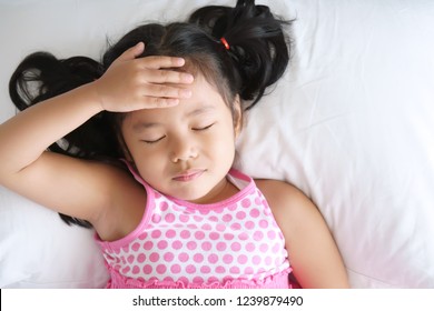 Asian Child Cute Or Kid Girl Sleep Headache Sick From Virus And Hand To The Forehead With Sad Or Cry On White Pillow And Bed With Wear Pink Dress At Home Or Children Hospital