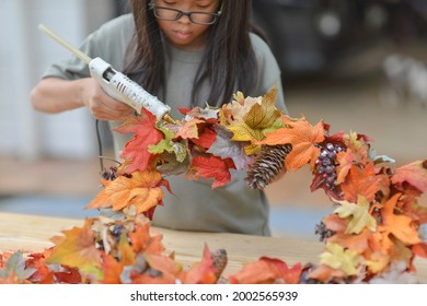 Asian Child Doing Fall Leaves Activity.Girl Holding Glue Gun To Make DIY Autumn Maple Wreath In The Outdoors. Young Female With Children Season School Project. Student With Creative Halloween Ideas.