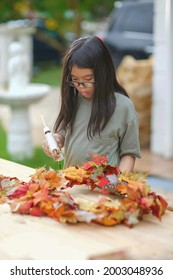 Asian Child Doing Fall Leaves Activity.Girl Holding Glue Gun To Make DIY Autumn Maple Wreath In The Outdoors. Young Female With Children Season School Project. Student With Creative Halloween Ideas.