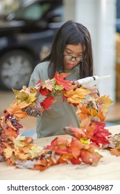 Asian Child Doing Fall Leaves Activity.Girl Holding Glue Gun To Make DIY Autumn Maple Wreath In The Outdoors. Young Female With Children Season School Project. Student With Creative Halloween Ideas.