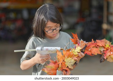 Asian Child Doing Fall Leaves Activity.Girl Holding Glue Gun To Make DIY Autumn Maple Wreath In The Outdoors. Young Female With Children Season School Project. Student With Creative Halloween Ideas.