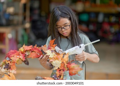 Asian Child Doing Fall Leaves Activity.Girl Holding Glue Gun To Make DIY Autumn Maple Wreath In The Outdoors. Young Female With Children Season School Project. Student With Creative Halloween Ideas.