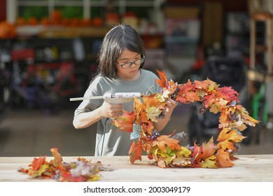 Asian Child Doing Fall Leaves Activity.Girl Holding Glue Gun To Make DIY Autumn Maple Wreath In The Outdoors. Young Female With Children Season School Project. Student With Creative Halloween Ideas.