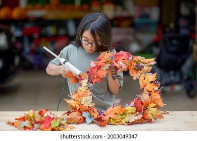 Asian Child Doing Fall Leaves Activity.Girl Holding Glue Gun To Make DIY Autumn Maple Wreath In The Outdoors. Young Female With Children Season School Project. Student With Creative Halloween Ideas.