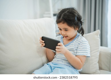 Asian child girl looking using and touch mobile phone screen on couch sofa. Baby smiling funny time to use mobile phone. Too much screen time. Cute girl watching videos while tv, Internet addiction. - Powered by Shutterstock