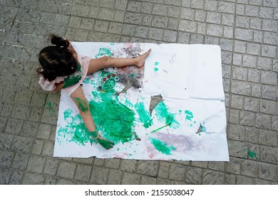 Asian Child Girl Sitting On Rip Canvas On Tile Floor In The Garden.Painted Green Color On Hands So Dirty, Leave A Mark On White Paper. Children's Creativity.Overhead Shot Of Painting Process. Child Dr