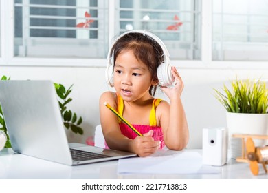 Asian Child Girl Studying Video Conference Distant Education At Home. Little Kid Preschool Wear Headphones Sit At Desk Use Laptop Computer And Communicates On Internet Online Video Call With Teacher